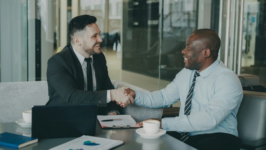 Two businessmen seated across a table in a modern office or cafe setting, shaking hands and smiling, symbolizing a successful agreement. The table holds items like a laptop, documents, coffee cups, and a clipboard with papers. The background features large glass panels and a sleek, professional environment. The scene offers networking events Seattle as a backdrop to foster professional connections.