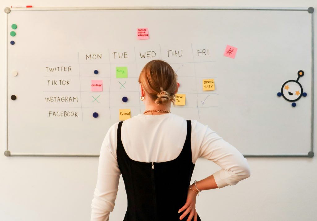 A person stands in front of a large whiteboard organized as a social media content calendar, a perfect example of the best way to keep up on social media trends. The board features a grid with days of the week as columns and social media platforms (Twitter, TikTok, Instagram, Facebook) as rows. Colored sticky notes with labels like 'Funny,' 'Culture,' and 'Product' mark planned content, while checkmarks, X's, and dots indicate task statuses. The person, wearing a black sleeveless top over a white shirt, observes the board with their hand on their hip. The scene conveys a structured and creative approach to social media marketing planning.