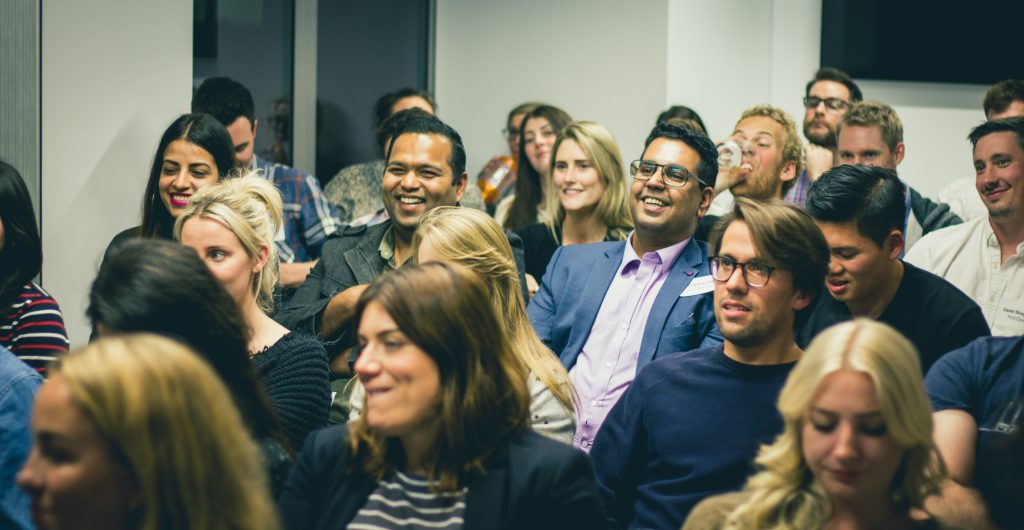 A diverse group of people sits in an audience or classroom setting, engaged and smiling as they look forward, likely at a speaker or presentation at one of the many networking events Tampa has to offer. The foreground features individuals slightly out of focus, including a woman with dark hair and a striped shirt. The background shows more attendees and light gray walls, creating a professional and positive atmosphere, possibly at a conference or workshop.