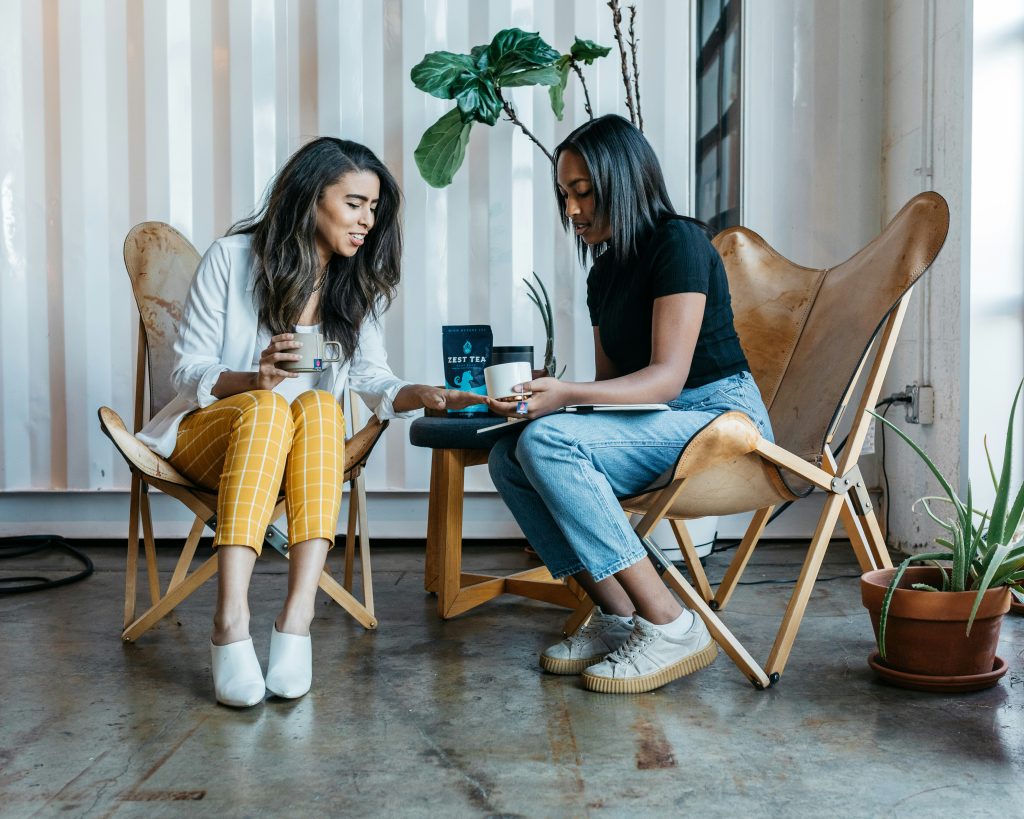 Two women seated in leather butterfly chairs, enjoying tea in a modern, industrial-style setting. A small table between them holds a blue pouch of tea and a black cylindrical object. One woman wears a white blouse and yellow plaid pants, holding a grey mug, while the other wears a black top and jeans, holding a white mug with a gold rim. The background features a corrugated wall, potted plants, and a polished concrete floor. The space offers networking events Seattle in a cozy and stylish environment.